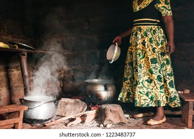 a black woman holding a ceramcic bowl standing in front of a large pot over an open flame.